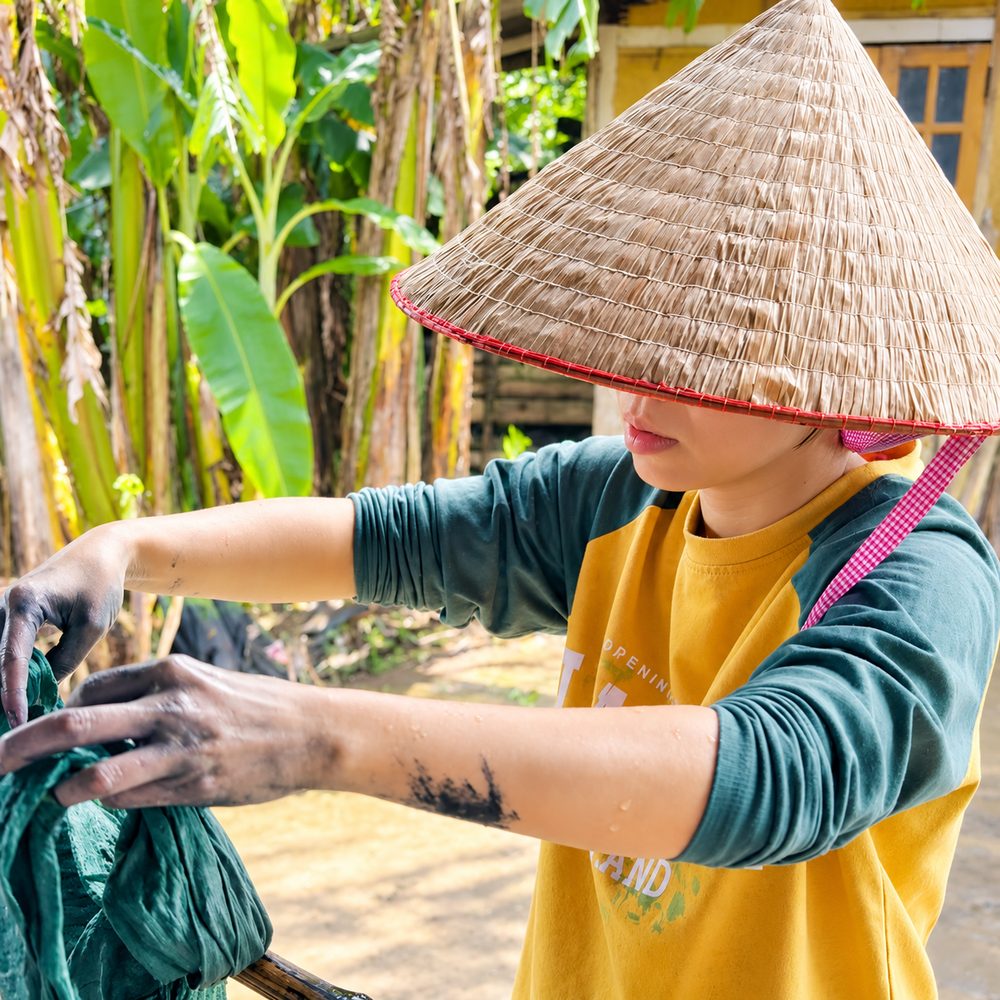 Tiên đội nón lá, hai tay nhuốm màu chàm đang vắt tấm vải vừa nhuộm bên vườn chuối